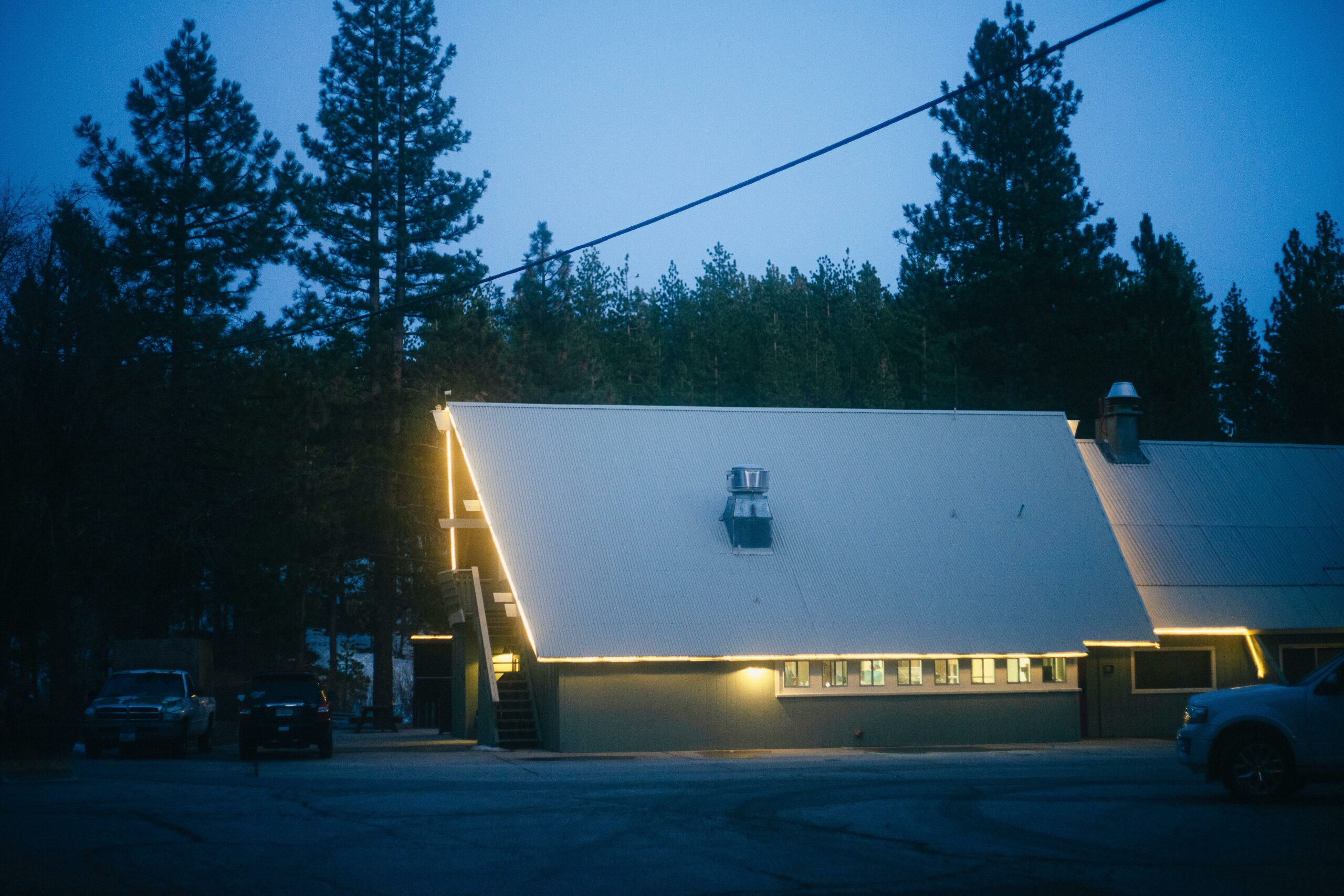 A large building with a steep A-frame roof is lit with warm, glowing lights outlining its structure. The building is surrounded by tall pine trees and a clear, darkening sky. Several vehicles are parked near the building, and the overall scene gives a cozy, inviting ambiance.