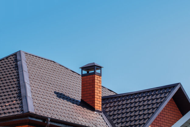 A modern rooftop with dark gray metal tiles and a red brick chimney topped with a black cap. The clean lines and bright blue sky create a sleek and minimalistic architectural appearance.