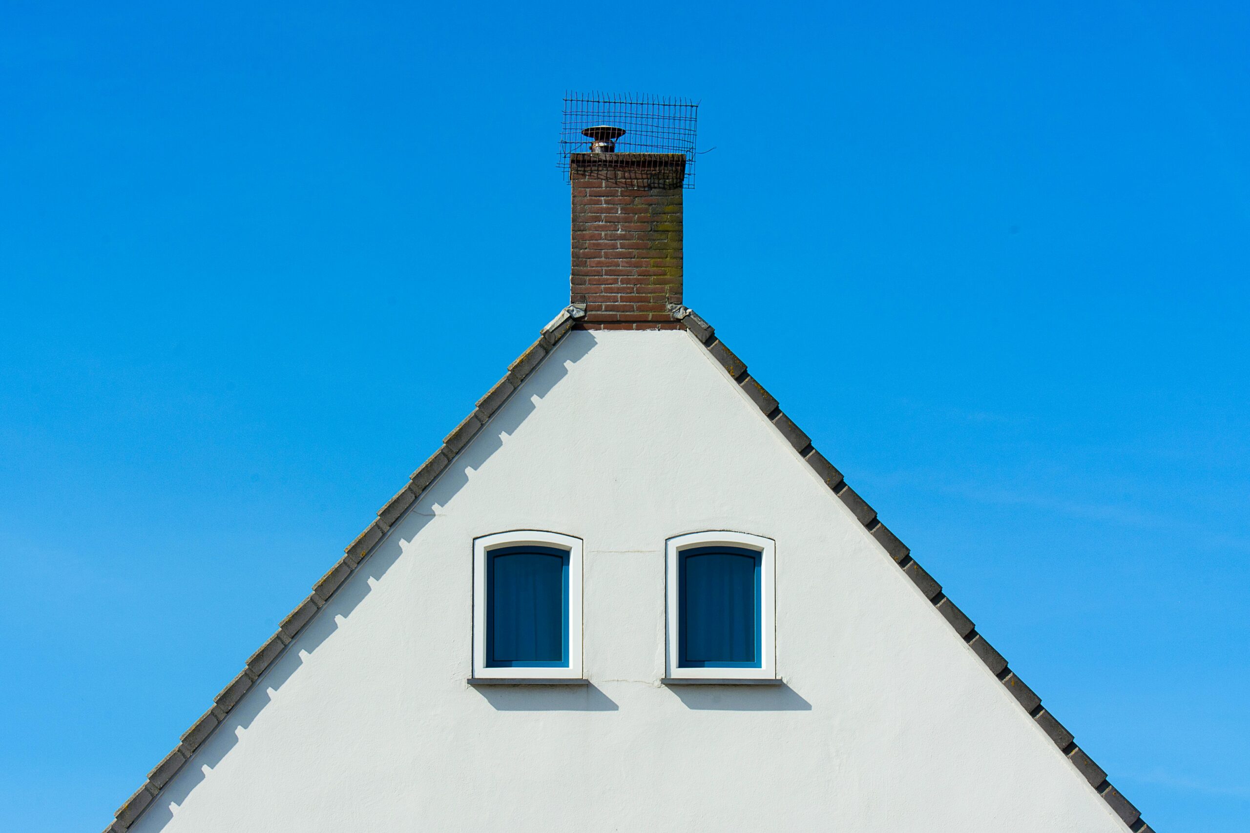 A white triangular gable of a house with two arched windows and a brick chimney at the top, covered with a protective metal mesh, set against a bright blue sky.